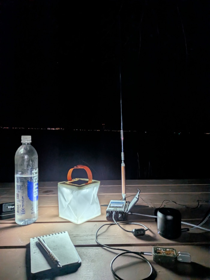 My KH1 and its whip antenna lit by lantern on a picnic table, with the Boston Harbor shoreline lights across the dark water