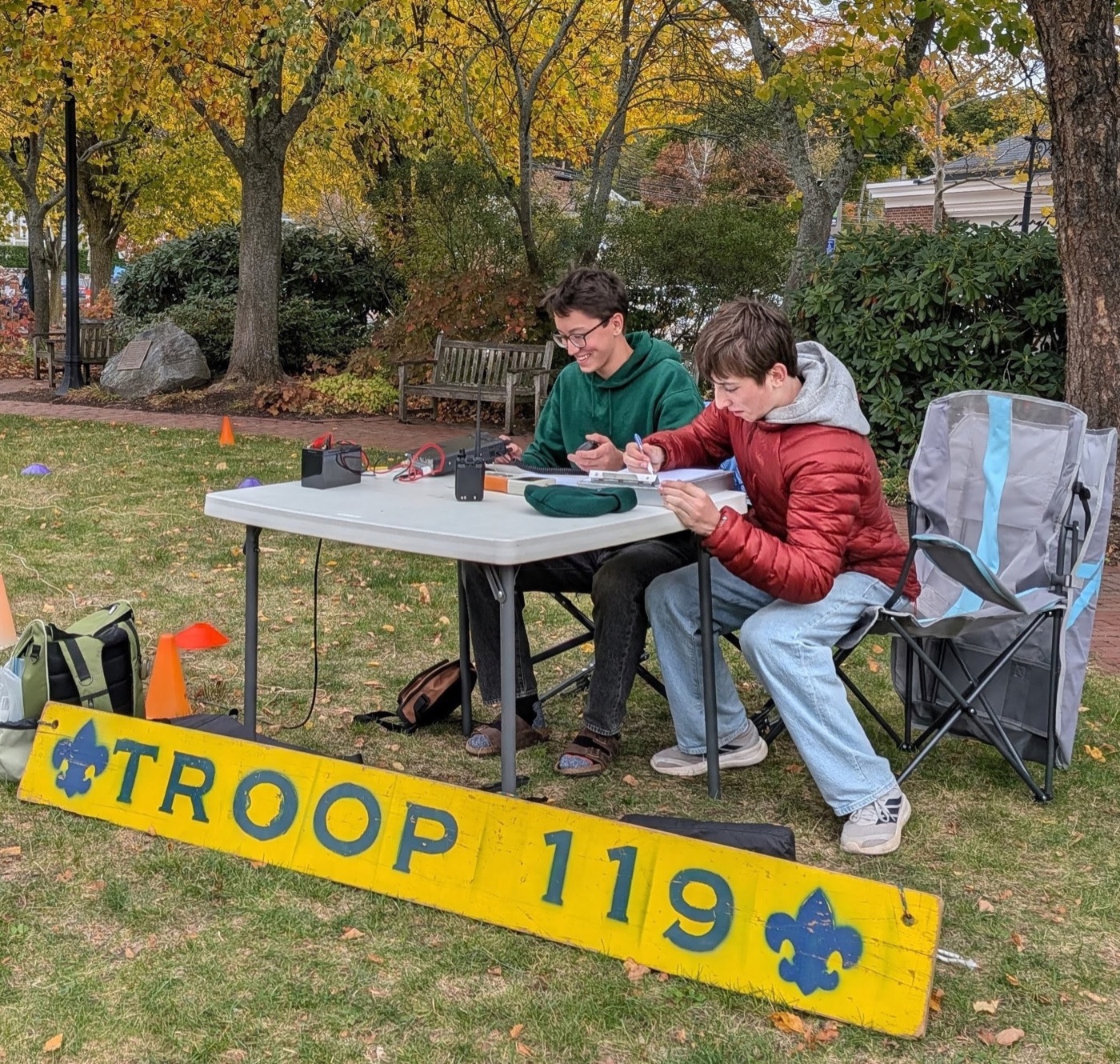 Two Scouts operating a radio at a folding table on a lawn in the town center, Tate (K1MKD) on the left talking on the microphone and another Scout logging, with a yellow Troop 119 sign in front