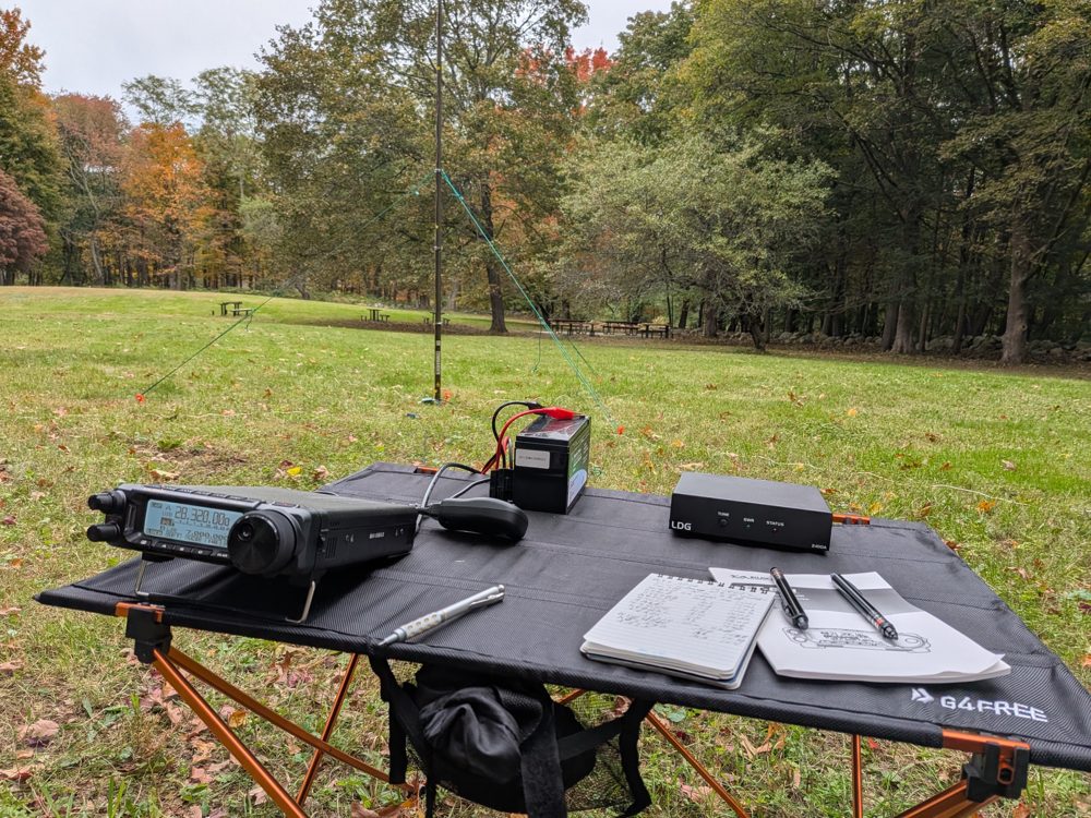 My FT-891, battery, LDG Z-100A tuner and notebook on a folding table with the Rybakov vertical standing in the field behind them