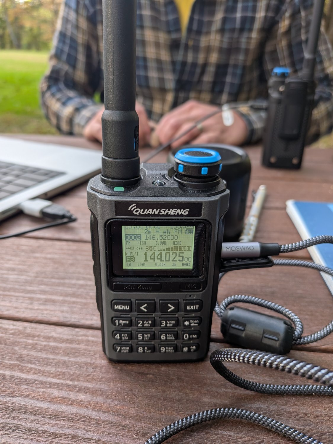 A Quansheng handheld radio on the picnic table, display showing 144.025 MHz in CW mode